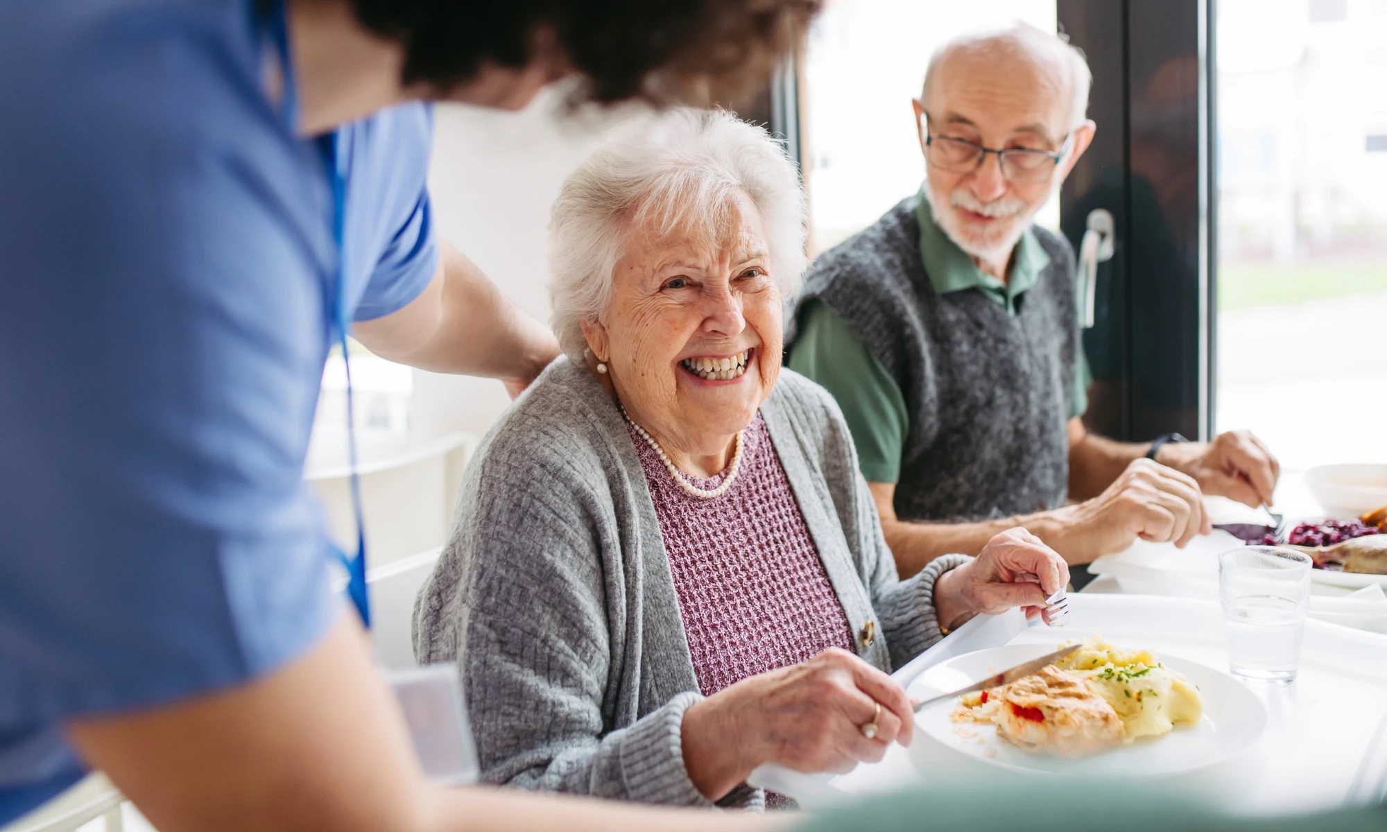 Caregiver serving lunch to a smiling senior living resident at the dining table